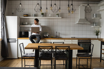 Freelancer working from home sitting on a kitchen worktop and using laptop. Bearded man working with a laptop and reading news. Handsome successful self entrepreneur working at his modern home.