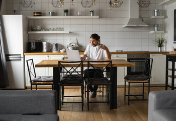 Freelancer working from home on the kitchen and using laptop. Handsome smiling man holding cup of coffee and looking at laptop. Successful self entrepreneur sitting with laptop and working at his