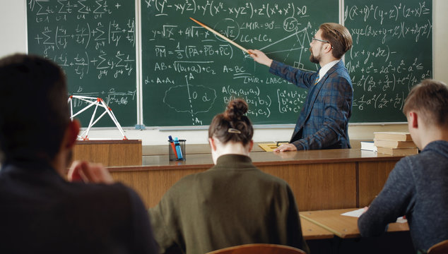 Math teacher in classic suit pointing at green chalkboard giving explanations to students