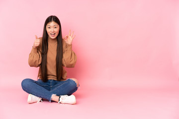 Young asian woman sitting on the floor isolated on pink background showing ok sign and thumb up gesture