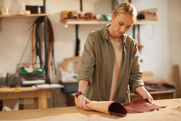 Young woman wearing casual clothes standing at table in her workshop rolling out piece of brown leather material
