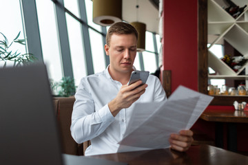 Young businessman wearing a white fashion shirt chatting and talking business on a mobile cell phone in a cafe with a laptop and documents. Freelance and selfemployment concept. Distance dream job.