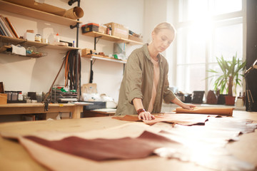Young woman standing at table in her workshop rolling out leather material for new craftwork, horizontal shot