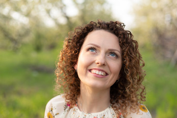 Close up portrait of young smiling attractive woman with curly hair in green flowering spring park. Pure emotions.