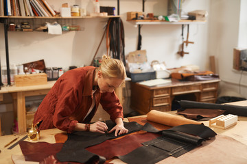 Fototapeta na wymiar Horizontal shot of young Caucasian craftswoman standing at table in workshop punching holes in black leather material parts to put rivets