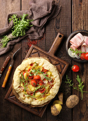 Concept of French and Italian cuisine. Pie with vegetables and bacon. Potatoes, cherry tomatoes, onions, bacon, arugula and spices on an old wooden background. Background image, copy space