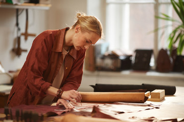 Horizontal side view medium shot of young Caucasian female artisan cutting out leather for new...