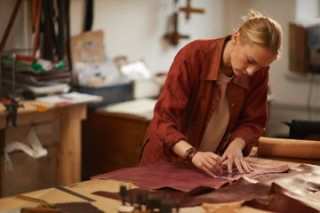 Horizontal medium portrait of concentrated young woman standing at table cutting out piece of leather material