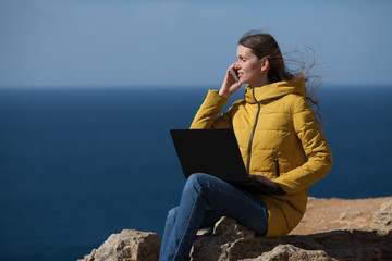 A freelancer girl is working typing on a laptop and talking on a mobile phone with a beautiful view of the open air sea sky. Traveling with a computer. Online dream job.