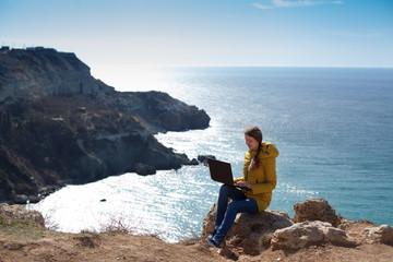 Technology and travel. Work in the open air. The concept of freelancing. A pretty young woman in a yellow jacket uses a laptop with a view of the sea Bay.