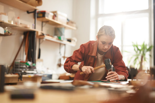 horizontal portrsit of young adult female artisan using hammer for leatherwork in modern workshop