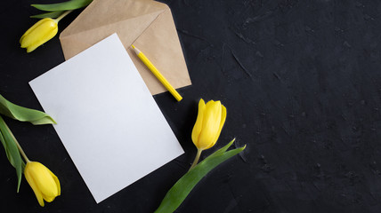 Yellow tulips on the edge of white paper for text on a dark background. A blank sheet of paper with an envelope. Copy space