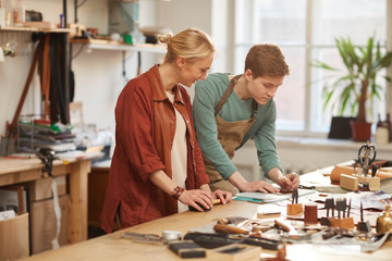 Horizontal shot of young man and cheerful woman working together in modern leather craft shop
