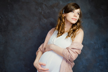 Pregnant young woman touching her belly. Beautiful expectant mom on dark background. Pregnancy, maternity clothes concept.