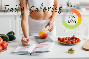 Cropped view of fit sportswoman writing calories while weighing apple on kitchen table, daily calories illustration - obrazy, fototapety, plakaty