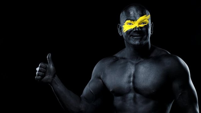 Man sports fan and bodybuilder athlete with yellow color on face art and black body paint. Colorful portrait of the guy with bodyart. Guy showing thumbs up sign with fingers.