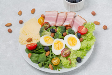 Plate with keto diet food. Keto breakfast: eggs, cheese, brisket, lettuce, broccoli, spinach, arugula, almonds, hazelnuts, blueberries and tomatoes.