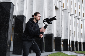 Young Professional videographer holding professional camera on 3-axis gimbal stabilizer. Pro equipment helps to make high quality video without shaking. Cameraman wearing black hoodie making a videos. © Volodymyr_sh