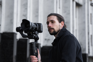 Young Professional videographer holding professional camera on 3-axis gimbal stabilizer. Pro equipment helps to make high quality video without shaking. Cameraman wearing black hoodie making a videos.