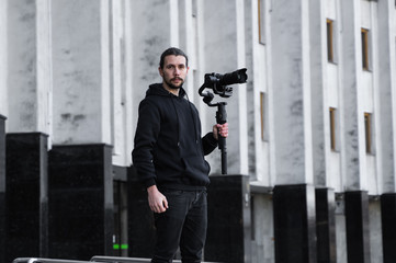 Young Professional videographer holding professional camera on 3-axis gimbal stabilizer. Pro equipment helps to make high quality video without shaking. Cameraman wearing black hoodie making a videos. © Volodymyr_sh