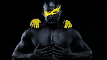 Man with female hands on the body. Bodybuilder athlete with yellow face art and black body paint. Colorful portrait of the guy with bodyart.