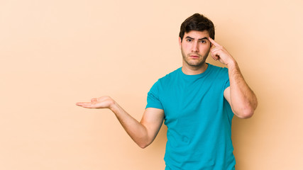 Young man isolated on beige background holding and showing a product on hand.