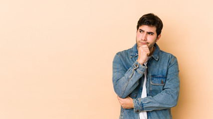 Young man isolated on beige background looking sideways with doubtful and skeptical expression.