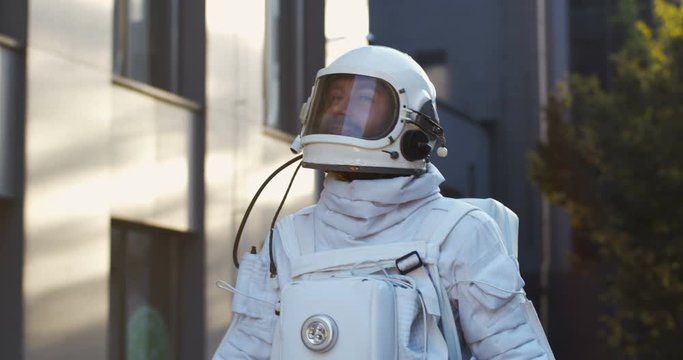 Close up of Caucasian male cosmonaut in space suit walking the town street and looking at building on sunny day. Spaceman in head armor in city. Coming back concept.