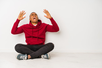 Young latin man sitting on the floor isolated screaming to the sky, looking up, frustrated.