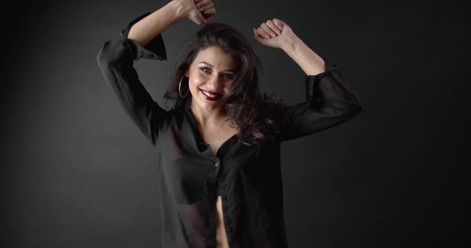 Happy stunning lady with long curly hair and bright makeup dancing while posing on camera with dark background. Professional model in black blouse enjoying working process.