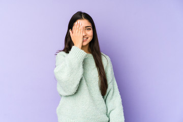 Young indian woman isolated on purple background having fun covering half of face with palm.