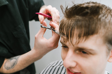 Background. Closeup photo of a barber clipping a happy blond client's hair scissors. Young man is hair cutg in a men's barber shop. Hairdresser trims young guy's bangs. Copy space