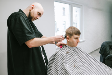Bald man professional barber cuts the client's hair with a clipper in the hands, looks at the young man's head. A young man cuts his barber. Barbershop concept.