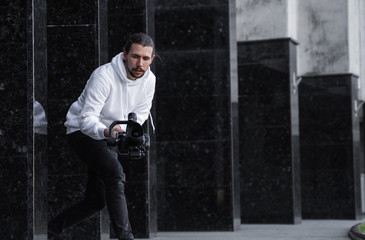 Young Professional videographer holding professional camera on 3-axis gimbal stabilizer. Pro equipment helps to make high quality video without shaking. Cameraman wearing white hoodie making a videos. © Volodymyr_sh