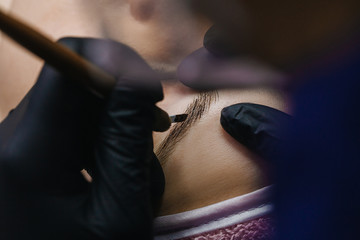 Hands in black gloves hold the manipulator with the microblading needle and draw her hair on the girl’s eyebrows close-up.