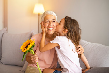 Little preschool granddaughter kissing happy older grandma on cheek giving violet flowers bouquet...