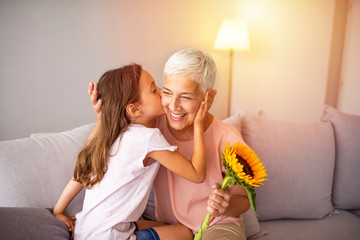 Cute girl giving a bunch of flowers to her grandmother sitting on the couch. Happy grandmother hugging small cute grandchild thanking for flowers presented. Darling granddaughter always remembers - obrazy, fototapety, plakaty