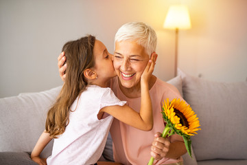 Cute girl giving a bunch of flowers to her grandmother sitting on the couch. Happy grandmother hugging small cute grandchild thanking for flowers presented. Darling granddaughter - obrazy, fototapety, plakaty