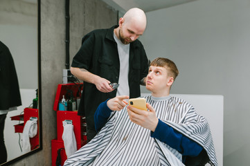 Young man client sits in a barber shop chair and shows a smartphone screen to his barber.Guy cuts his hair at a men's hairdresser,points finger at the screen of a smartphone and looks at a hairdresser