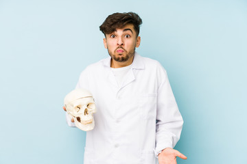 Young arabian doctor man holding a skull shrugs shoulders and open eyes confused.