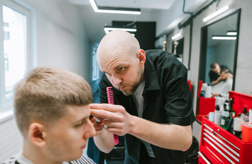 Professional barber is focused on trimming a young client's hair. Professional hairdresser does a hairstyle for a young man in a barbershop,looking with a serious face