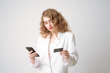 Confident young curly woman in white blazer holds credit card in one hand and phone in other, isolated over grey background
