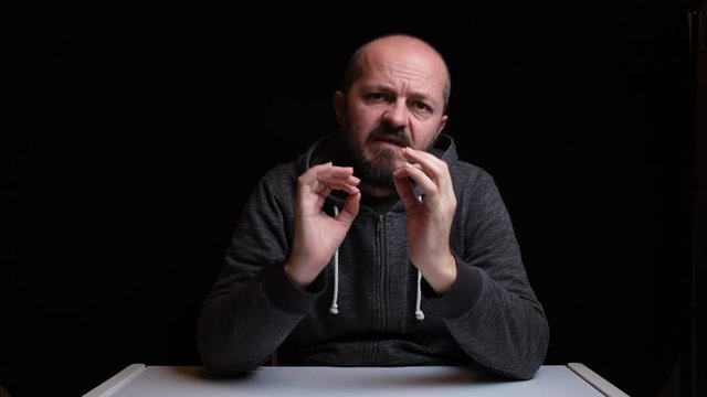 Studio footage of bearded Caucasian man in sweater sitting behind desk and talking to camera, isolated against black background, video blog concept