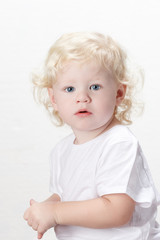 Naklejka premium Blond Curly Haired Caucasian Toddler Boy with Blue Eyes Wearing White Shirt Sitting Down Looking Unsure