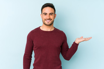 Young caucasian man isolated on blue background showing a copy space on a palm and holding another hand on waist.