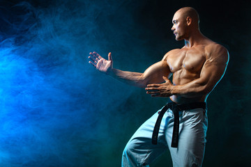 Fototapeta na wymiar Karate or taekwondo fighter on black background with smoke. Fit man sportsmen bodybuilder physique and athlete. Men's sport motivation.