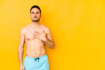 Young caucasian man with swimsuit isolated on yellow background Young caucasian man with trtaking an oath, putting hand on chest.
