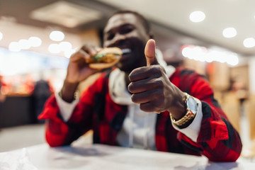 A portrait of young african man eating a burger in street food cafe. Fast food eating time. Thumb up finger.
