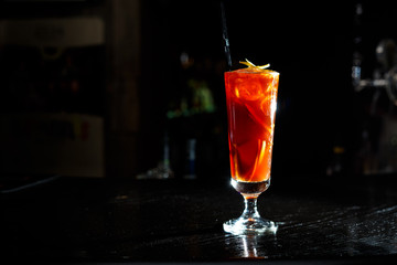 A red cocktail in a tall glass stands on the counter of a dark bar.