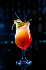 A red cocktail in a tall glass stands on the counter of a dark bar.